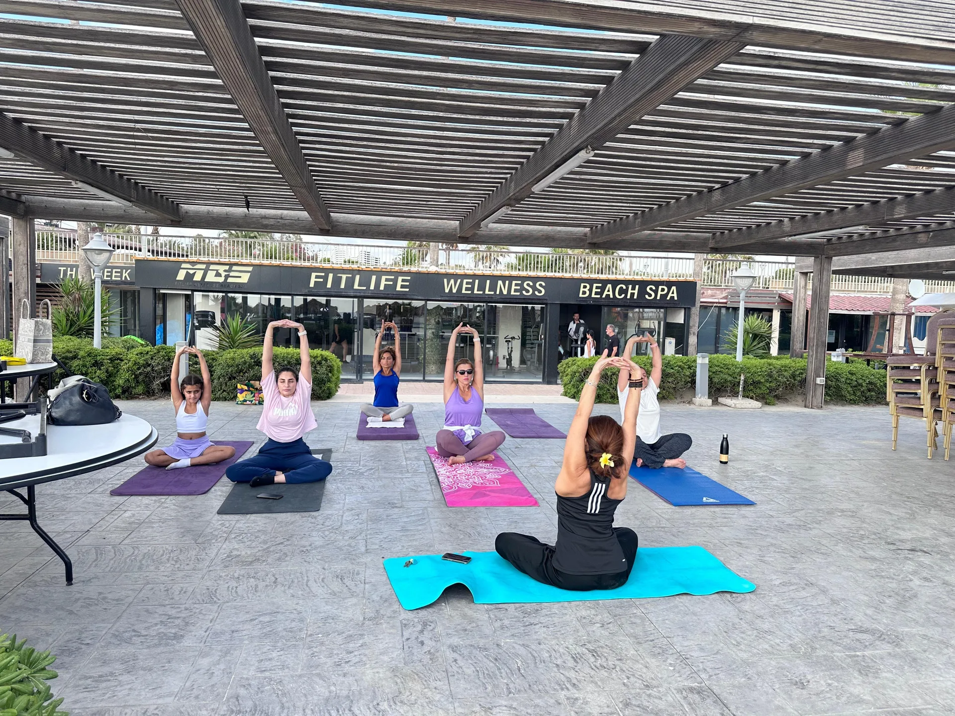 A beach yoga class stretching on mats under the pergola outside the studio